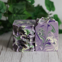 Load image into Gallery viewer, Two swirled purple and green soap bars on a wooden surface with a blurred green leafy background.