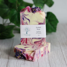 Load image into Gallery viewer, Two swirled soap bars with a visible brand label on a wooden surface with green leaves in the background.