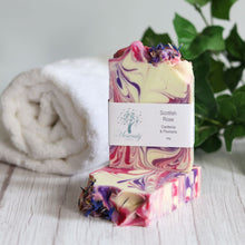 Load image into Gallery viewer, Marbled soap bar with a label on a wooden surface, towel, and plant in the background