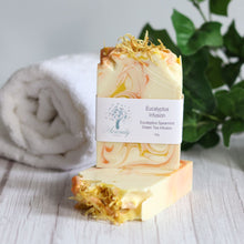 Load image into Gallery viewer, Bar of soap with a label on a wooden surface, towel, and plant in the background
