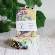 Load image into Gallery viewer, Colorful silk scarf with a label on a wooden surface with green leaves in the background
