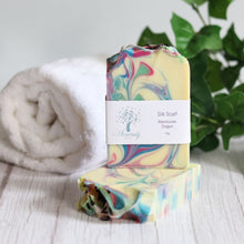 Load image into Gallery viewer, Colorful marbled silk scarf with a label on a wooden surface, towel, and plant in the background.
